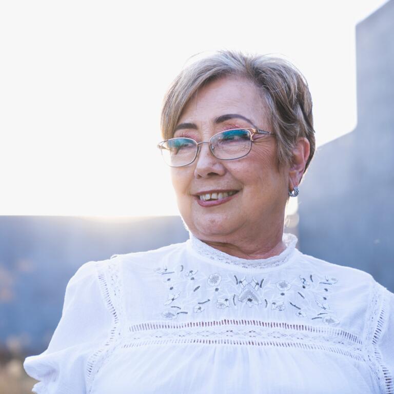 An elderly woman with short grey hair, glasses and a white blouse smiles.